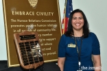 Sharmin Barolia stands in front of a plaque with the names of past honorees of the Embrace Civility Award.