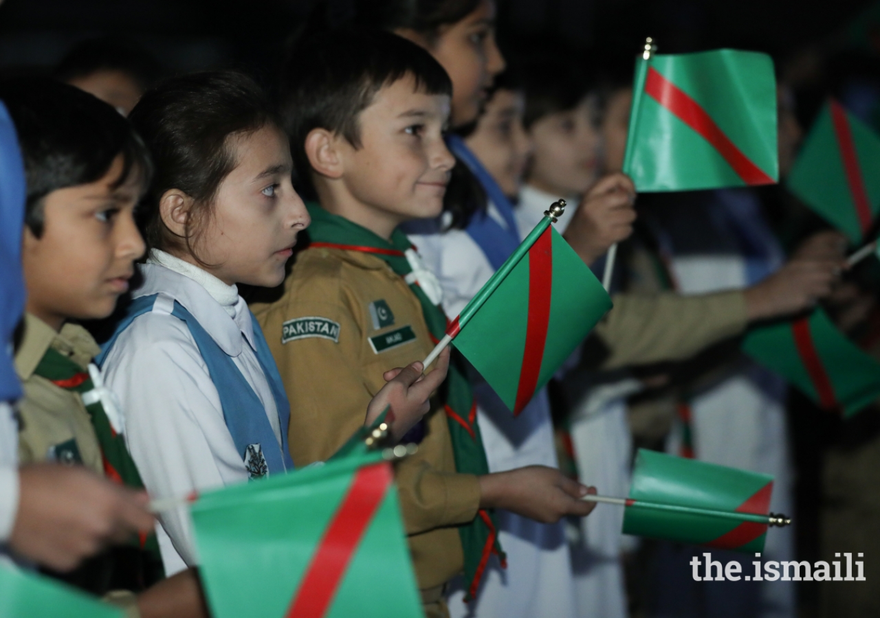 Young scouts and guides waving the national flag, the Ismaili flag and the Diamond Jubilee flag.