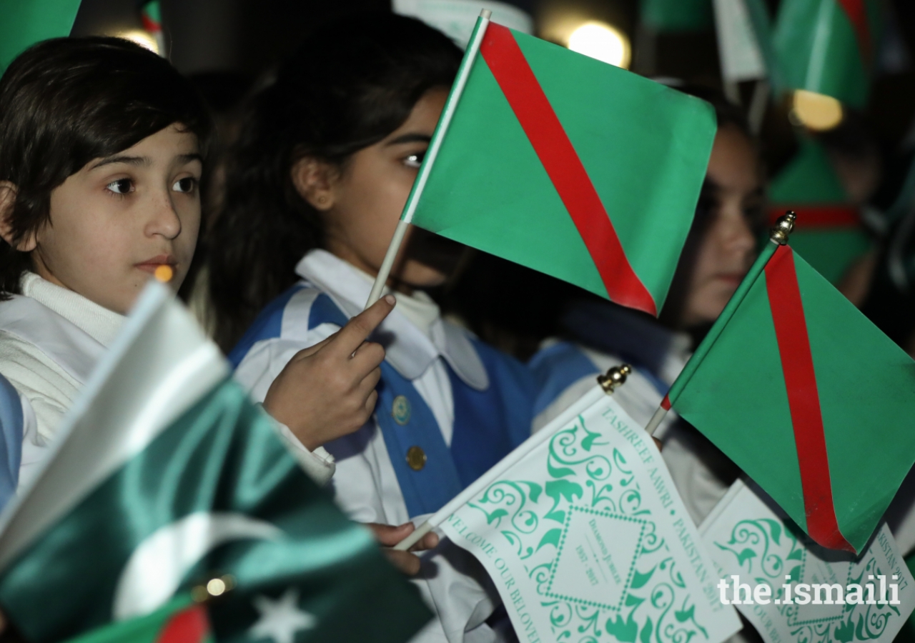 Young scouts and guides waving the national flag, the Ismaili flag and the Diamond Jubilee flag.